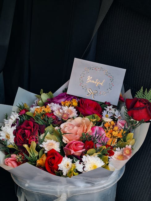 A vibrant floral bouquet arranged with a mix of red roses, peach lilies, pink roses, white daisies, and small yellow and white filler flowers, wrapped in white tissue paper with a clear cellophane out