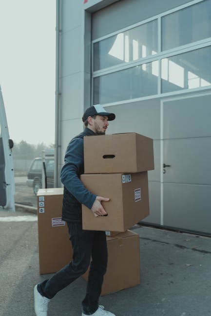 A delivery person holding three brown cardboard boxes outside a modern building with large glass windows, preparing for flower delivery service. The individual is dressed in a casual dark jacket, grey
