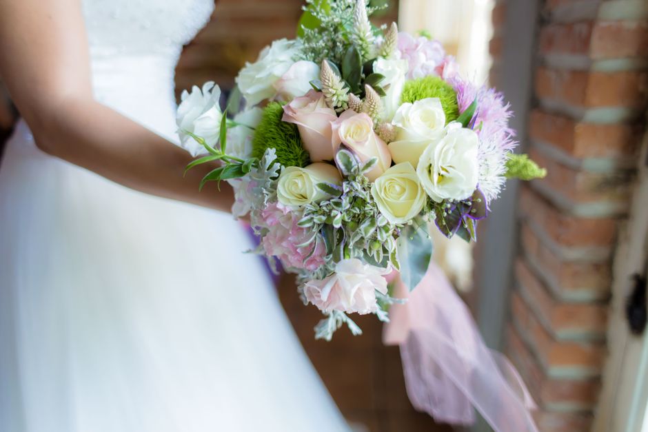 A close-up view of a bridal bouquet featuring fresh flowers, including white roses, pale pink lisianthus, and soft purple hyacinths, accented with green foliage and sprigs of dusty miller. The bouquet