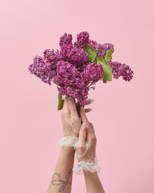 A pair of hands adorned with delicate lace gloves gently hold a fresh bouquet of purple lilacs with vibrant green leaves. The bouquet, composed of tightly clustered lilac flowers, showcases a soft, fr