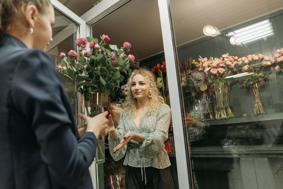 A woman with curly blonde hair, dressed in a light-colored floral blouse, is receiving a bouquet of pink roses wrapped in clear cellophane from a florist inside a flower shop. The florist, whose face 
