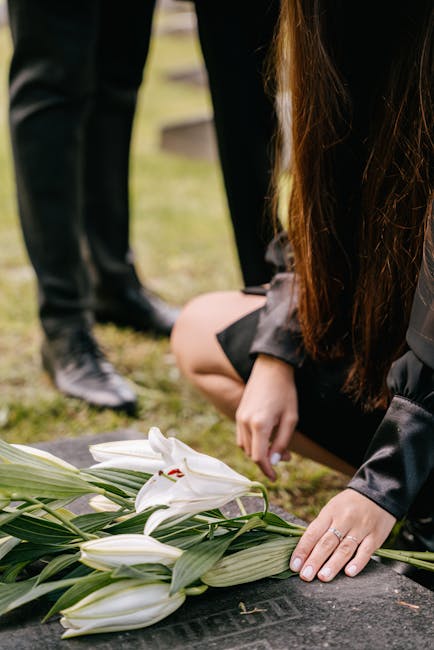 A person dressed in black, kneeling on the ground, arranging a bouquet of white lilies with long green leaves, possibly at a funeral or memorial setting. The floral arrangement is placed on a dark sur
