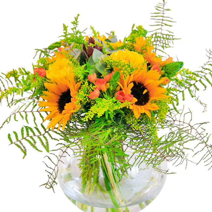 The first glance at this bouquet evokes pure sun-drenched delight, capturing the spirited heart of Elmers End. Large, bright sunflowers beam at the front, their petals layered in a dazzling array of yellows that conjure memories of golden afternoons in Alexandra Recreation Ground nearby. Beside them, buttery yellow garden roses open in generous, swirling formations, offering a classic romantic charm against the bold sunflowers. Deep green foliage and airy, vine-like greens flow outward, their movement hinting at the untamed vitality of local gardens in full bloom. Scarlet hypericum berries, polished to a gleam, glisten amongst the stems, providing dynamic contrast to the blooms. The bouquet is anchored in a perfectly round glass vase that reveals the clean lines of each submerged stem, grounding the vibrant colors in serene clarity. Subtle blue-grey eucalyptus leaves peek out, offering an aromatic freshness, while tiny yellow filler blossoms dance in the mix, reminding one of wildflowers along Brook Street. Up close, the tactile richness-fuzzy sunflower centers, plush rose petals, smooth greens-comes alive, mingled with hints of fragrance that evoke open fields. This arrangement suits any milestone: whether brightening a birthday table, celebrating an anniversary, or welcoming new beginnings in Elmers End, the flowers radiate true happiness and local charm.